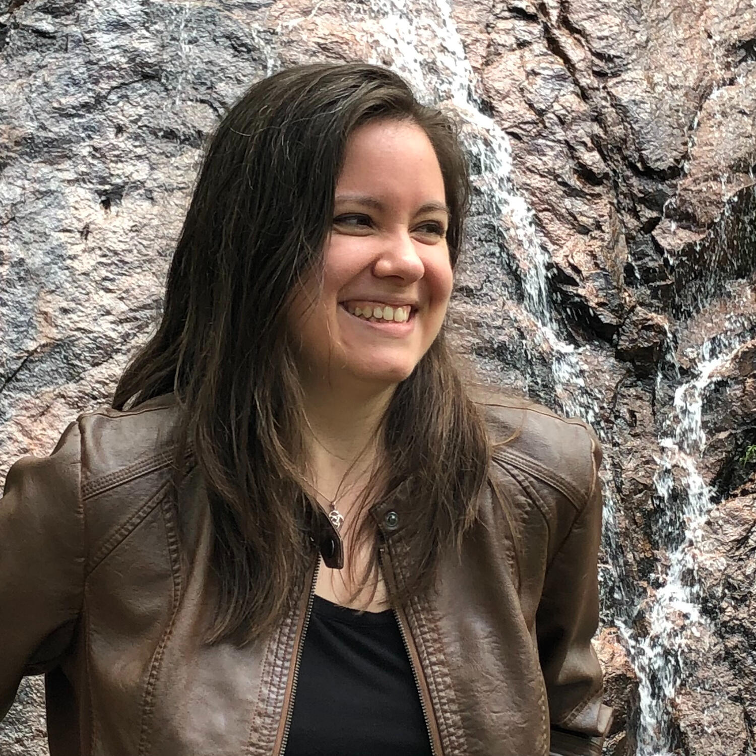 Kaila Desjardins A white woman with long brown hair and wearing a brown jacket smiling in front of a rock wall and small waterfall.